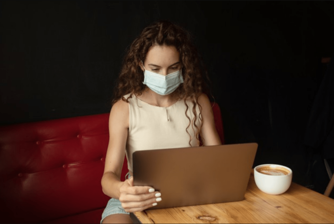 Chica joven con mascarilla usando su ordenador mientras se toma un café